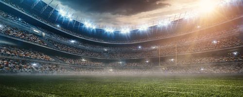 A wide angle panoramic image of a outdoor american football stadium full of spectators under evening sky. The image has depth of field with the focus on the foreground part of the pitch. The view from center of the field.